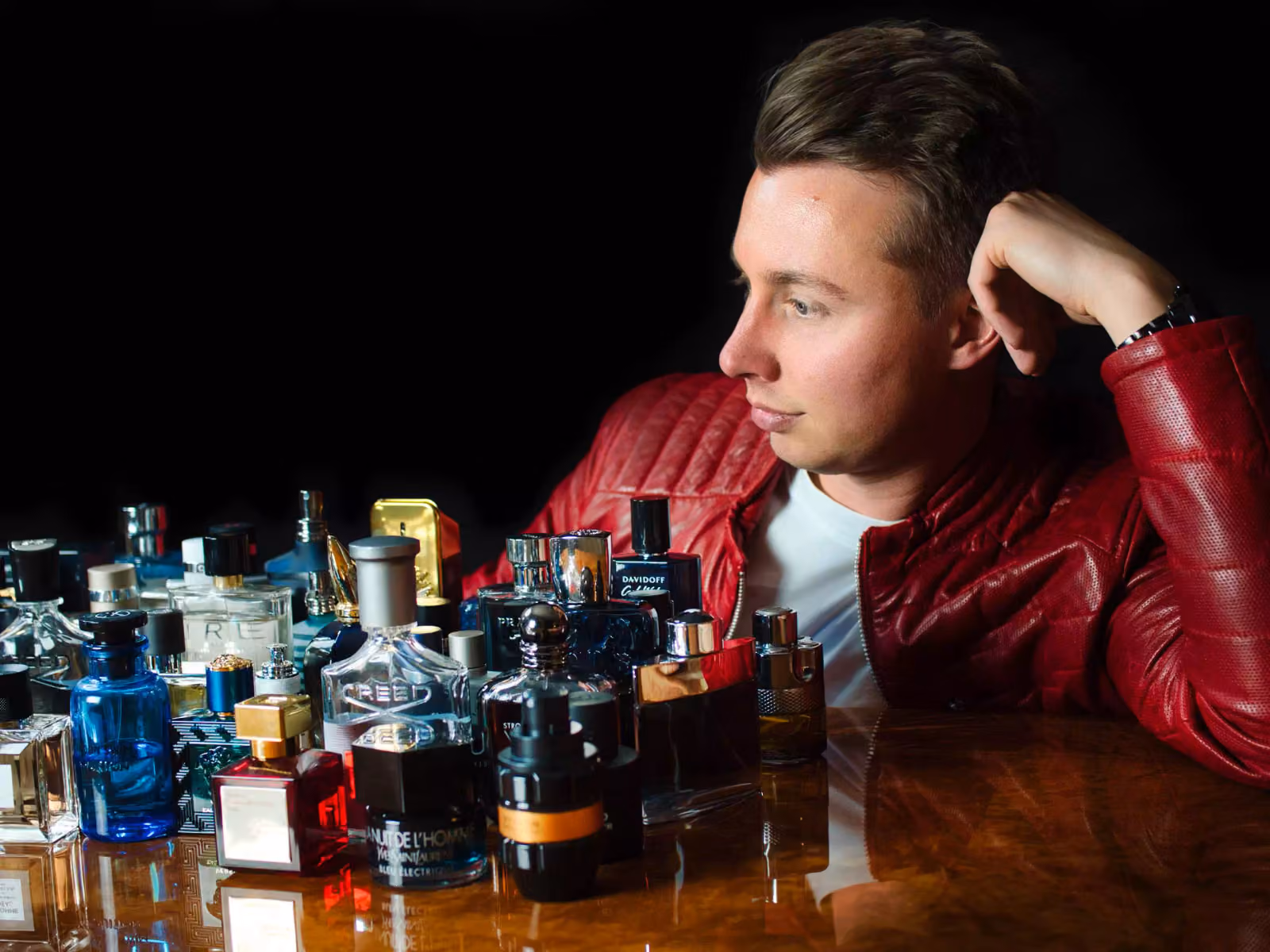 Peter Krück in a red leather jacket, resting his head on his hand and looking at a large collection of perfume bottles on a table